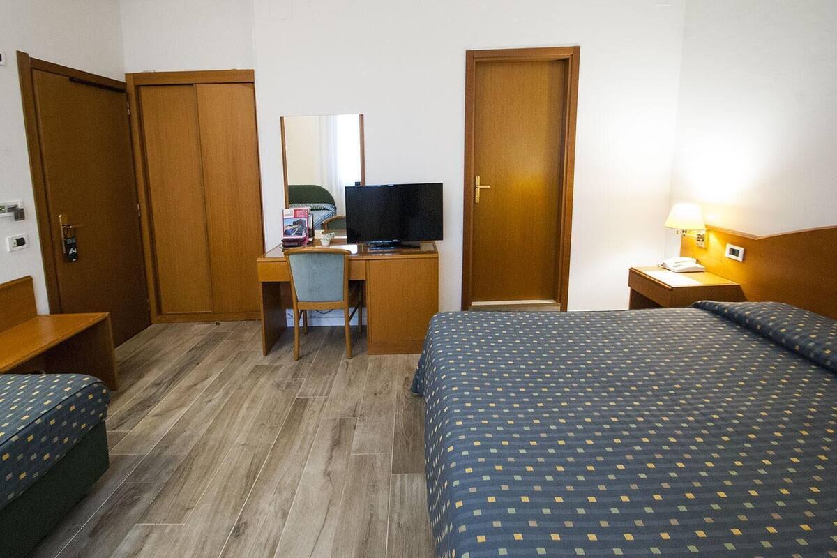 A hotel room with a bed, a desk with a chair, a television, and a bedside table with a lamp.