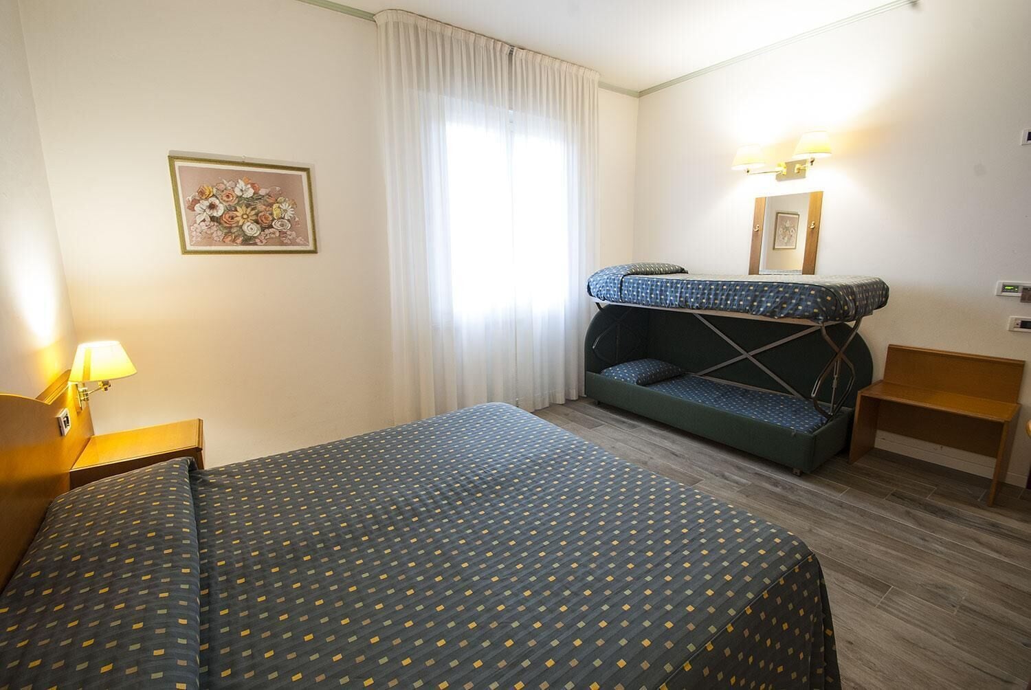 A hotel room with a bed, a sofa bed, a nightstand, a lamp, and a window with curtains.
