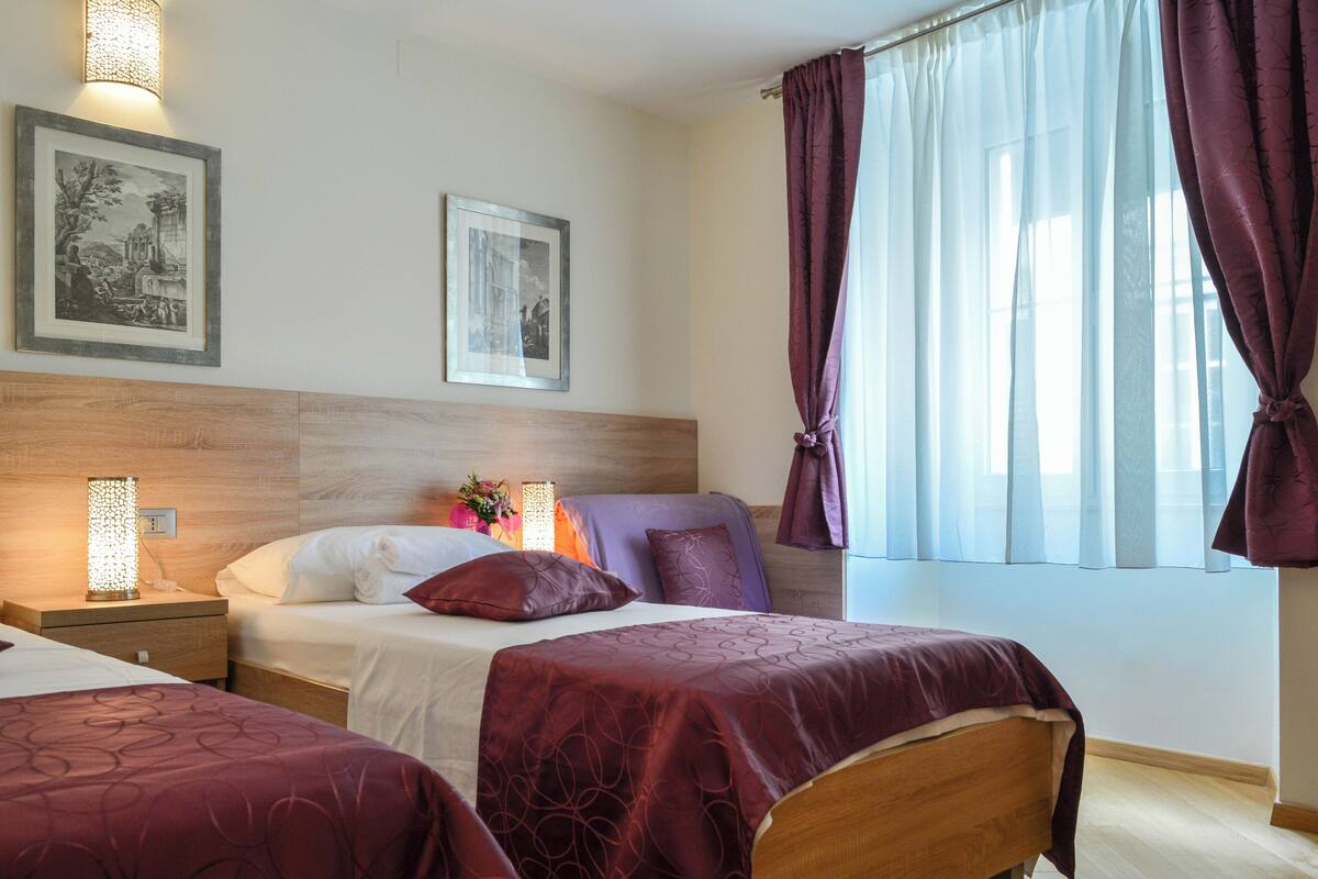 Hotellihuone, jossa on kaksi sänkyä, puinen pääty, seinällä kehystettyjä kuvia, yöpöytä lampun kanssa ja ikkuna, jossa on verhot.