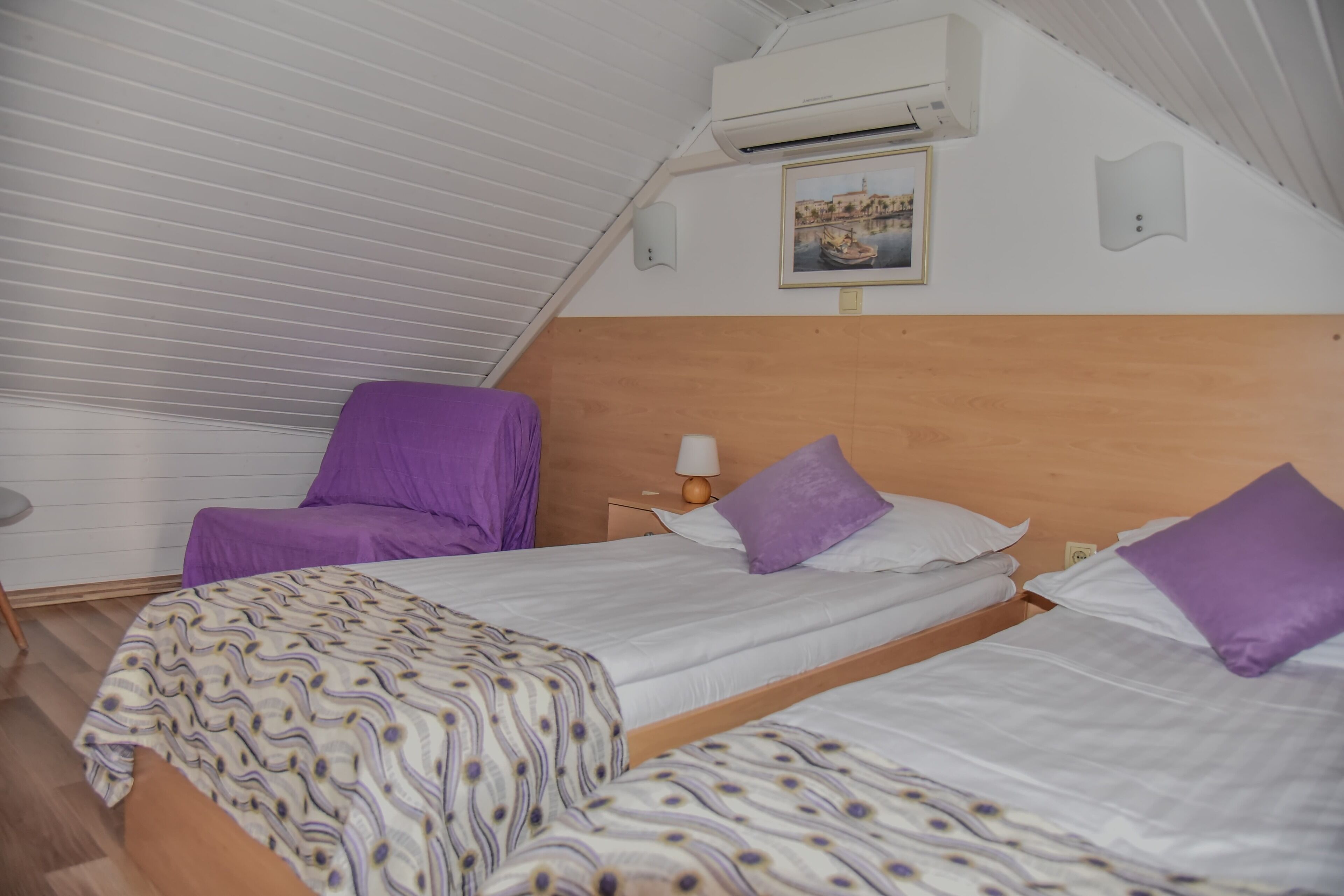 Een kamer met twee bedden, elk voorzien van een paars kussen en een bedsprei met een patroon. Er is een houten hoofdbord, een nachtkastje met een lamp en een airconditioner aan de muur.
