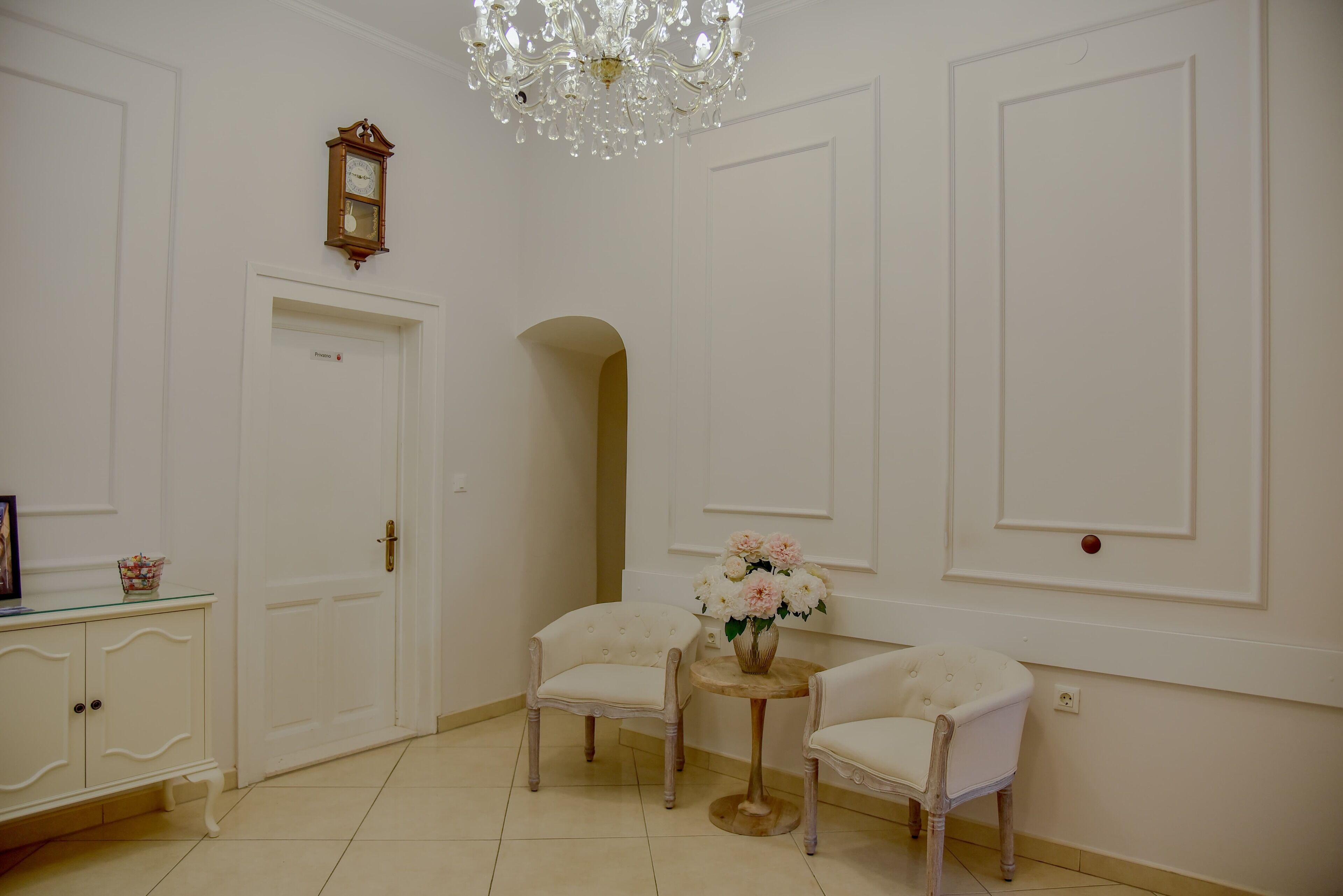 A room with white walls, a chandelier, a clock, two chairs, a small table with flowers, and a door.