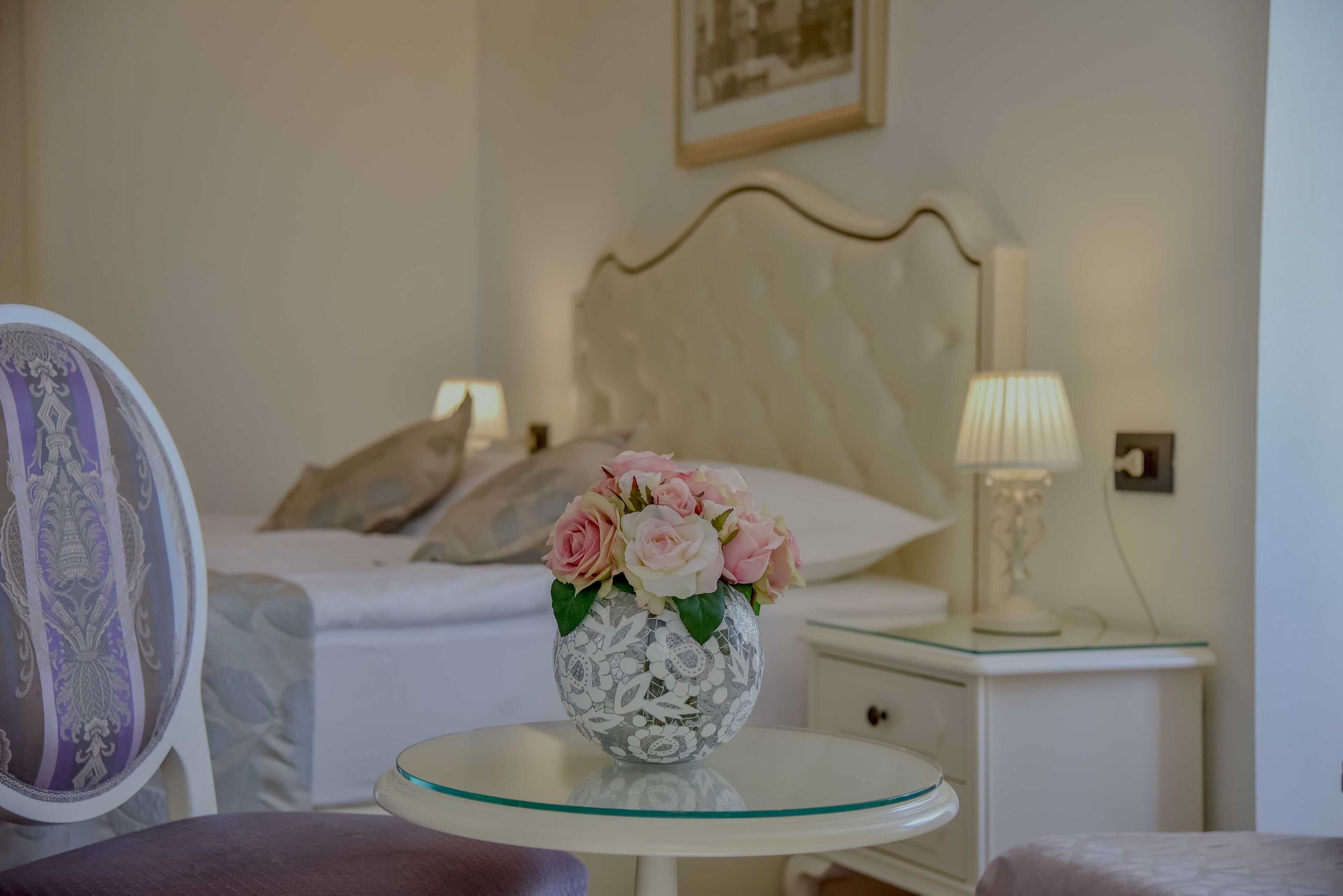 A bedroom with a bed, a bedside table with a lamp, and a vase with flowers on a small round table.