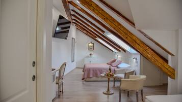 Ein Schlafzimmer mit geneigter Decke, Holzbalken, einem Bett mit rosa Bettdecke, einem Nachttisch mit Flasche und Gläsern sowie einem Stuhl.