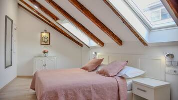 A bedroom with a sloped ceiling, wooden beams, a skylight, a bed with pink bedding, a bedside table, and a nightstand.