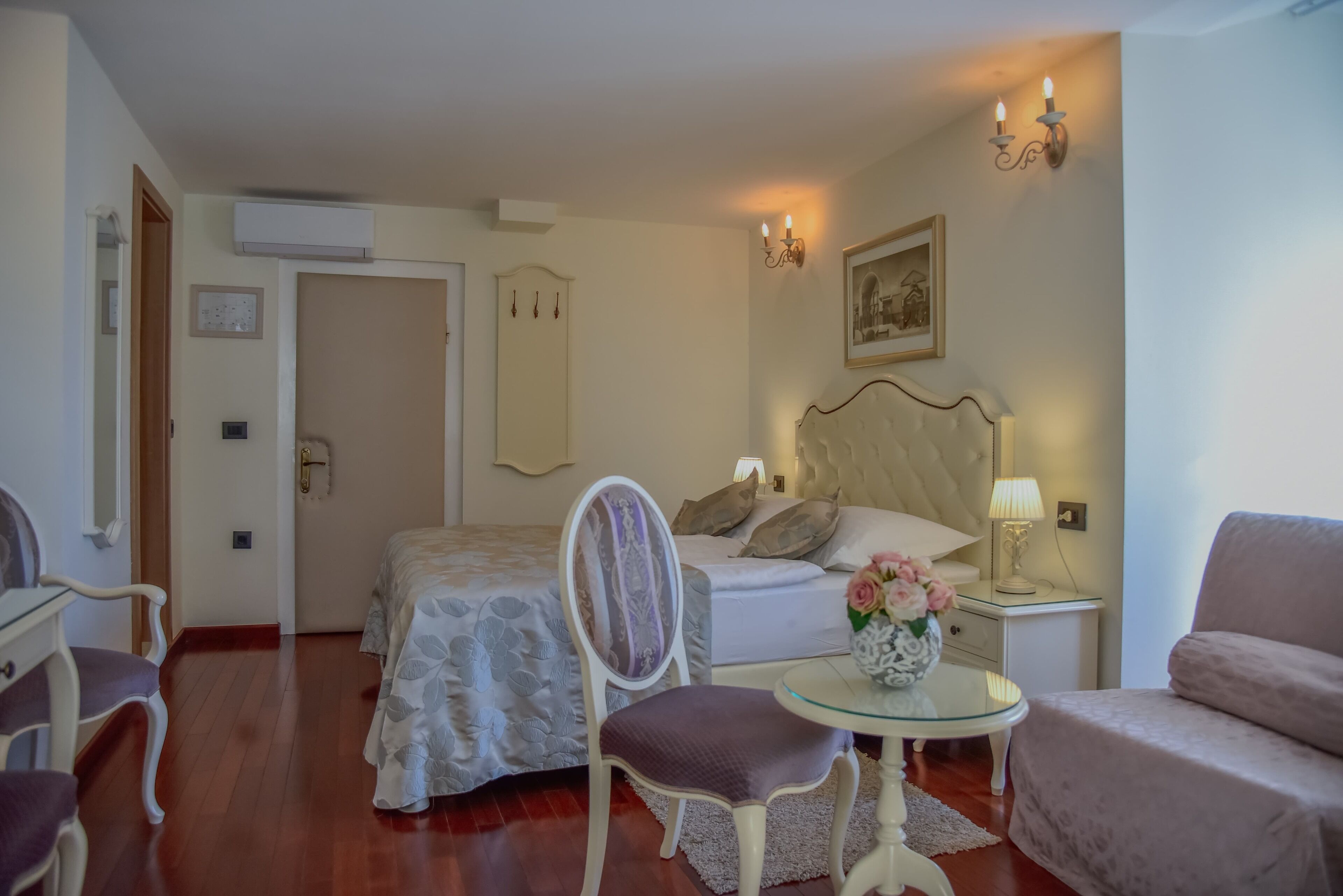 A hotel room with a bed, a chair, a small table with a vase, and a nightstand with a lamp.