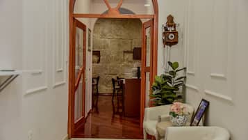 A hallway with a stone wall, wooden door frame, and a clock on the wall. There is a chair with a vase of flowers and a framed picture on a side table.