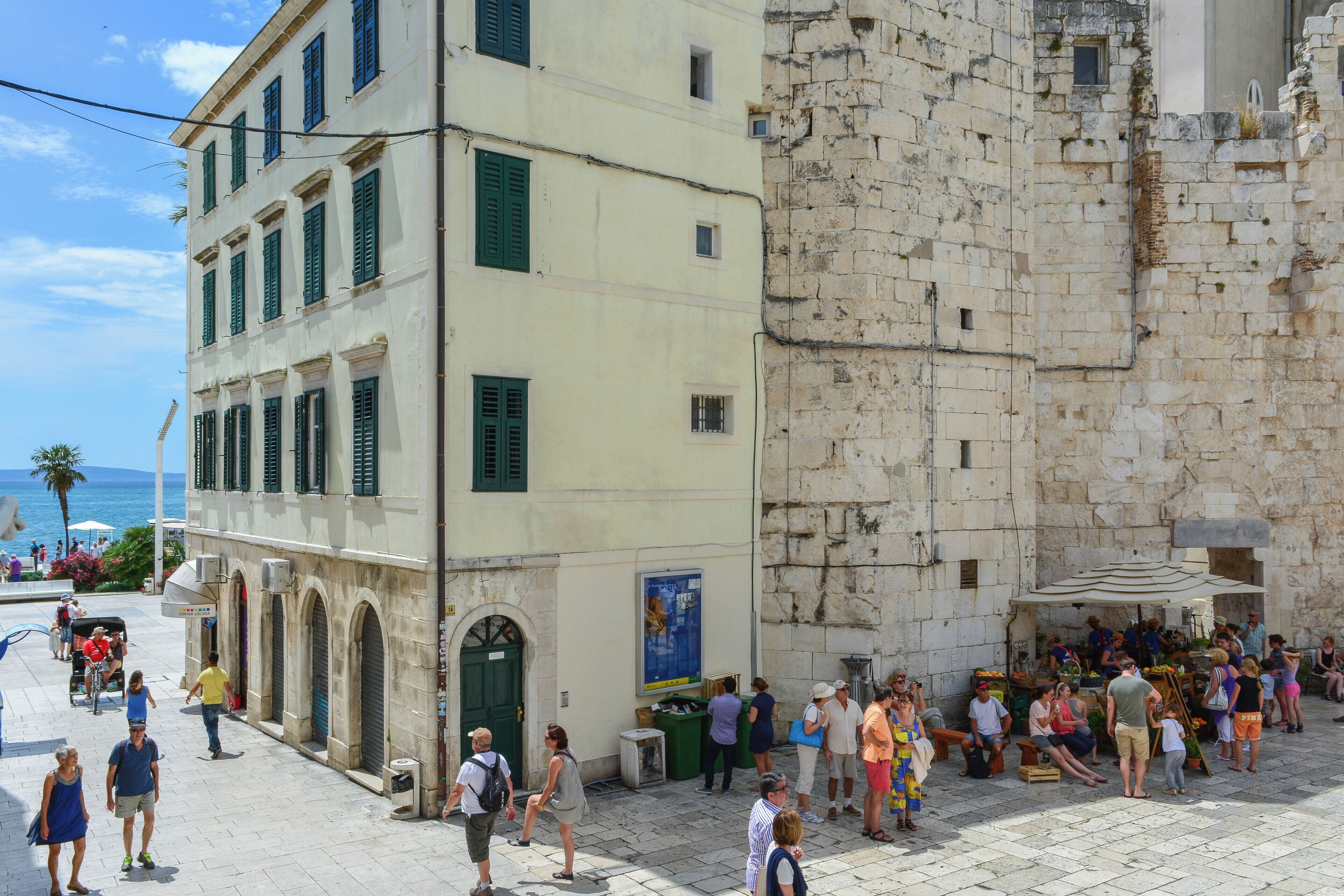 A bustling street scene with a mix of modern and historic architecture, including a building with green shutters and a stone wall.