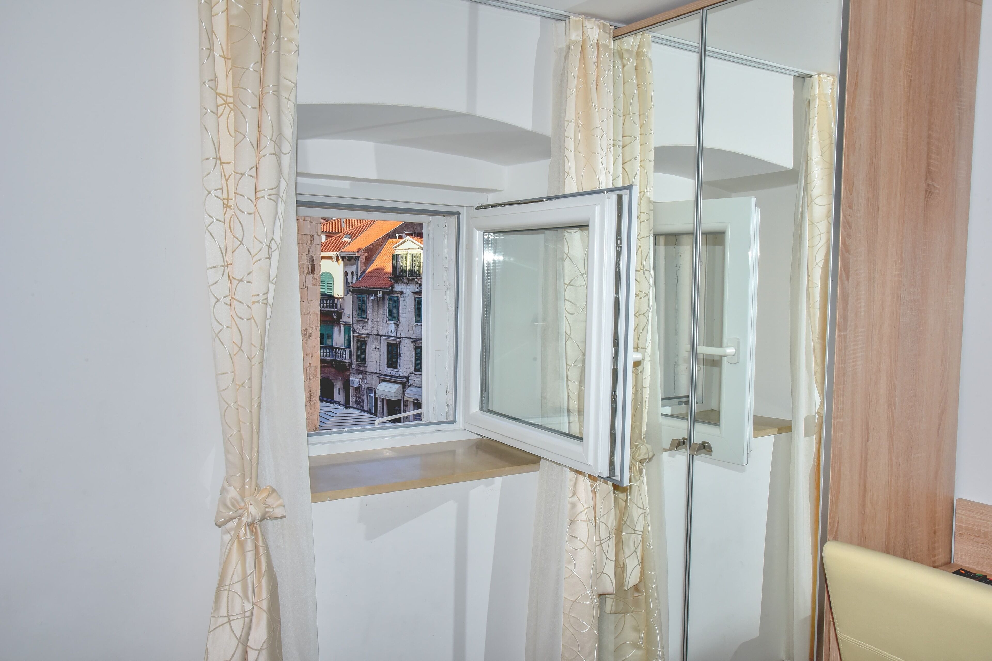 A room with a window, a mirror, and a view of buildings outside.