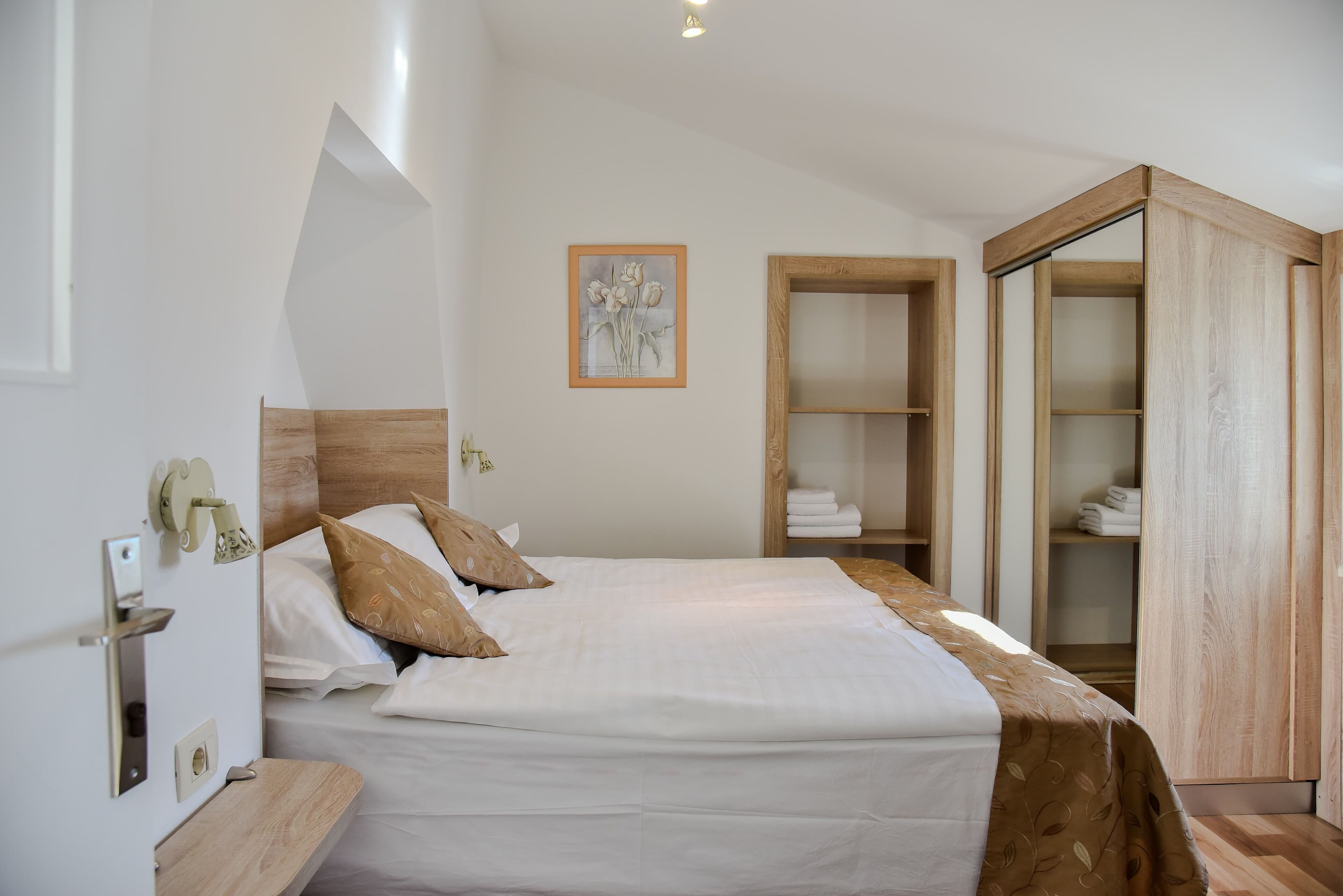 Een keurig opgemaakt bed met hoofdbord, een houten garderobe en een kunstwerk in een lijst aan de muur.