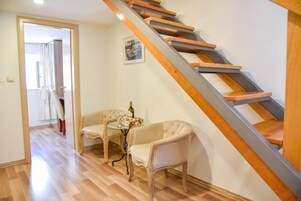A staircase with wooden steps and a metal handrail, leading to an upper floor. There is a small table with a bottle and glasses, and two chairs in the foreground.