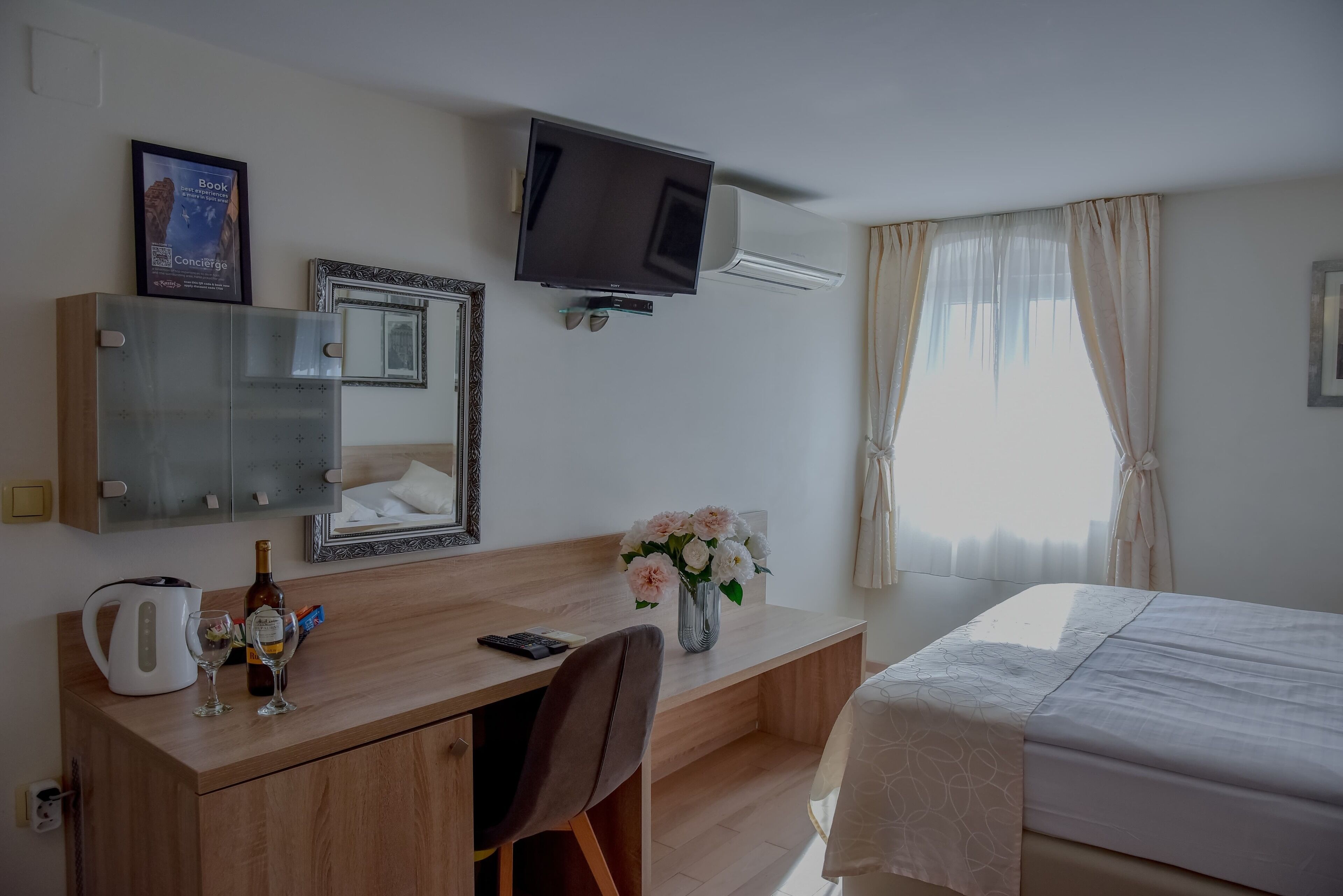 Una habitación de hotel con una cama, un escritorio con silla, un televisor en la pared, un espejo, un florero con flores y una ventana con cortinas.