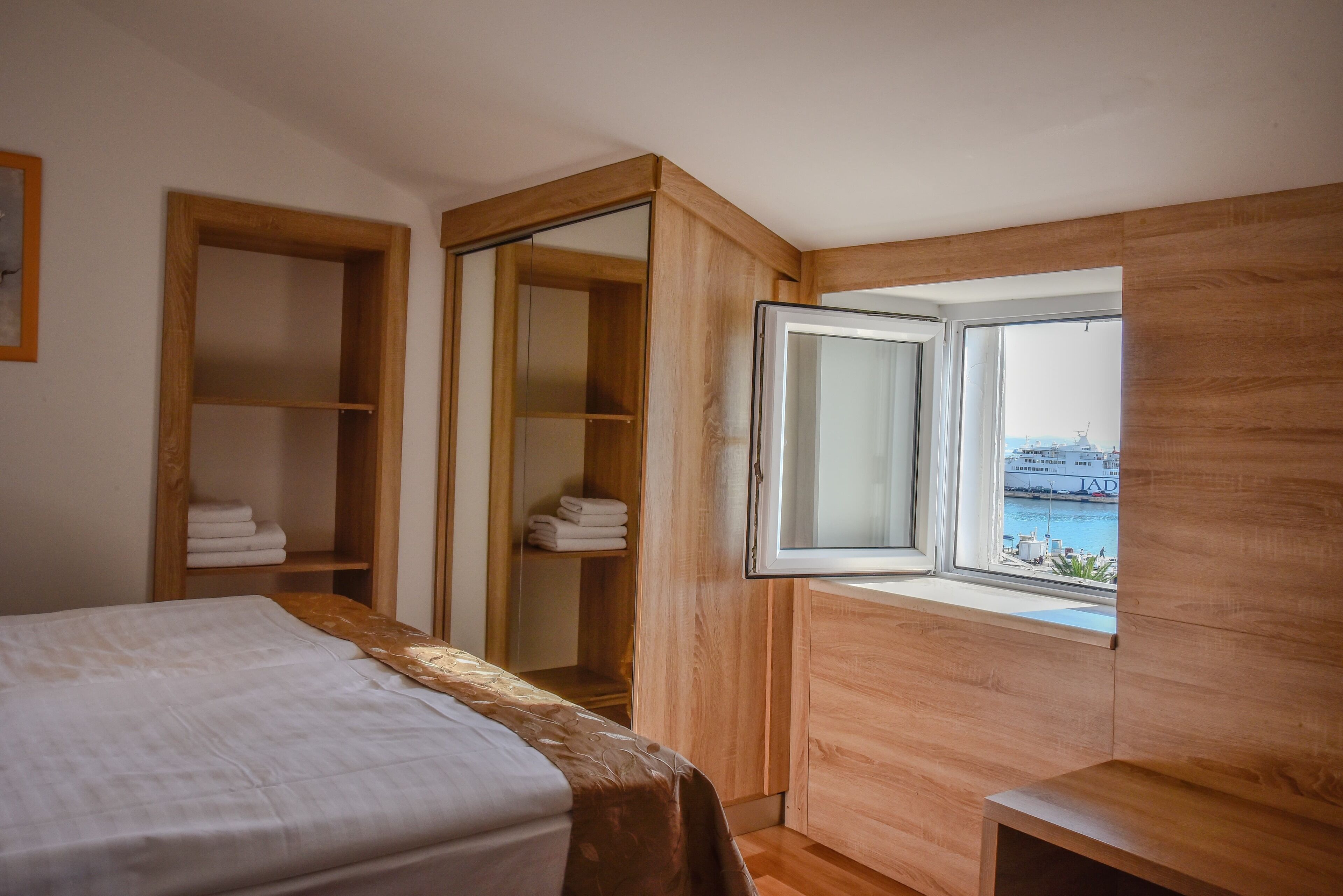 A bedroom with a bed, wooden wardrobe, and a window with a view of a ship.