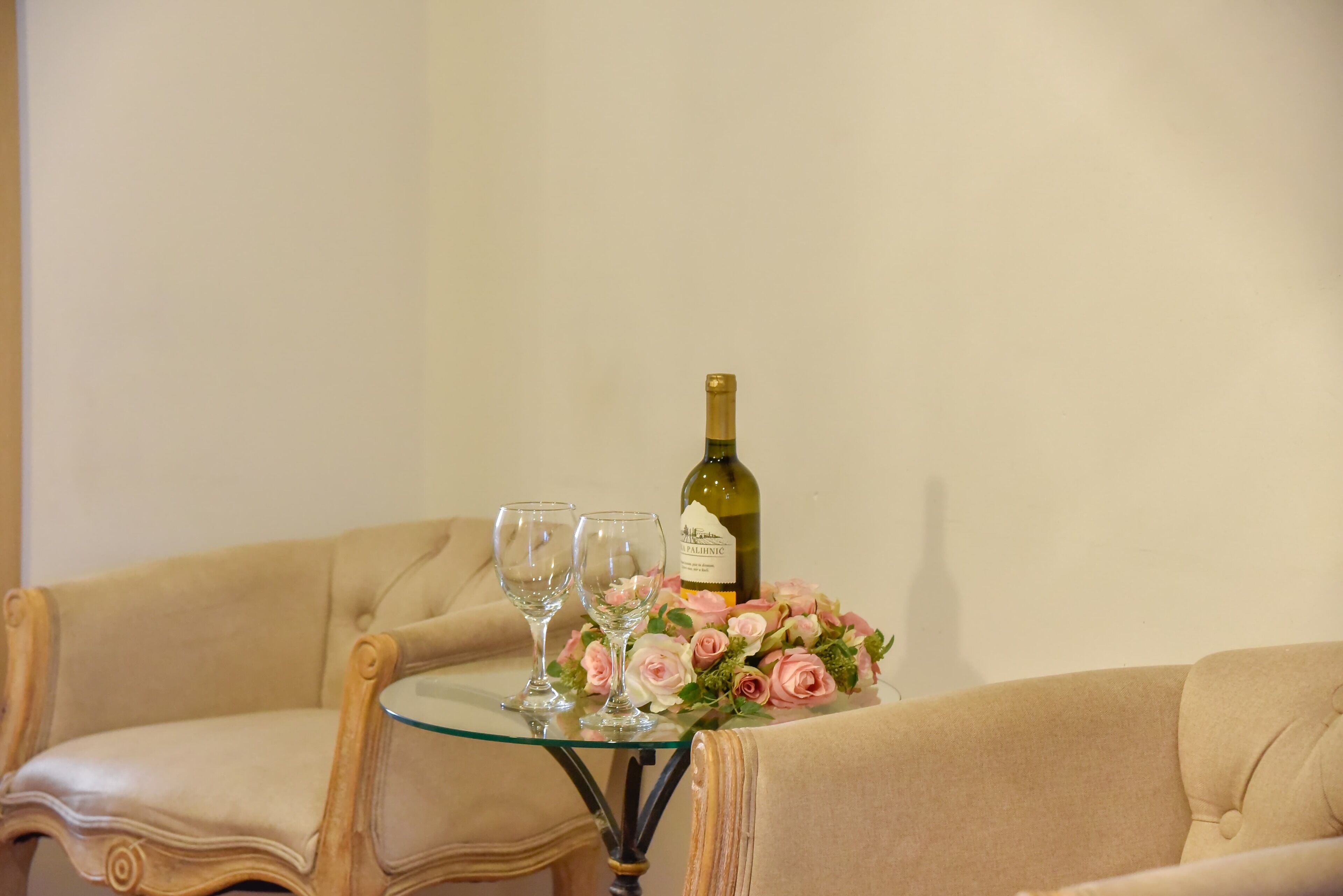 Et sofabord i glas med en vinflaske, to vinglas og en blomsterbuket på en beige sofa.