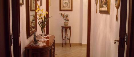 A hallway with a wooden dresser, a mirror, and a vase with yellow flowers. There are framed pictures on the walls and a small table with a plant in the center.
