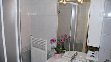 Une salle de bain avec un lavabo blanc, un miroir et une douche. On y trouve un porte-serviettes et une orchidée en pot sur le lavabo.