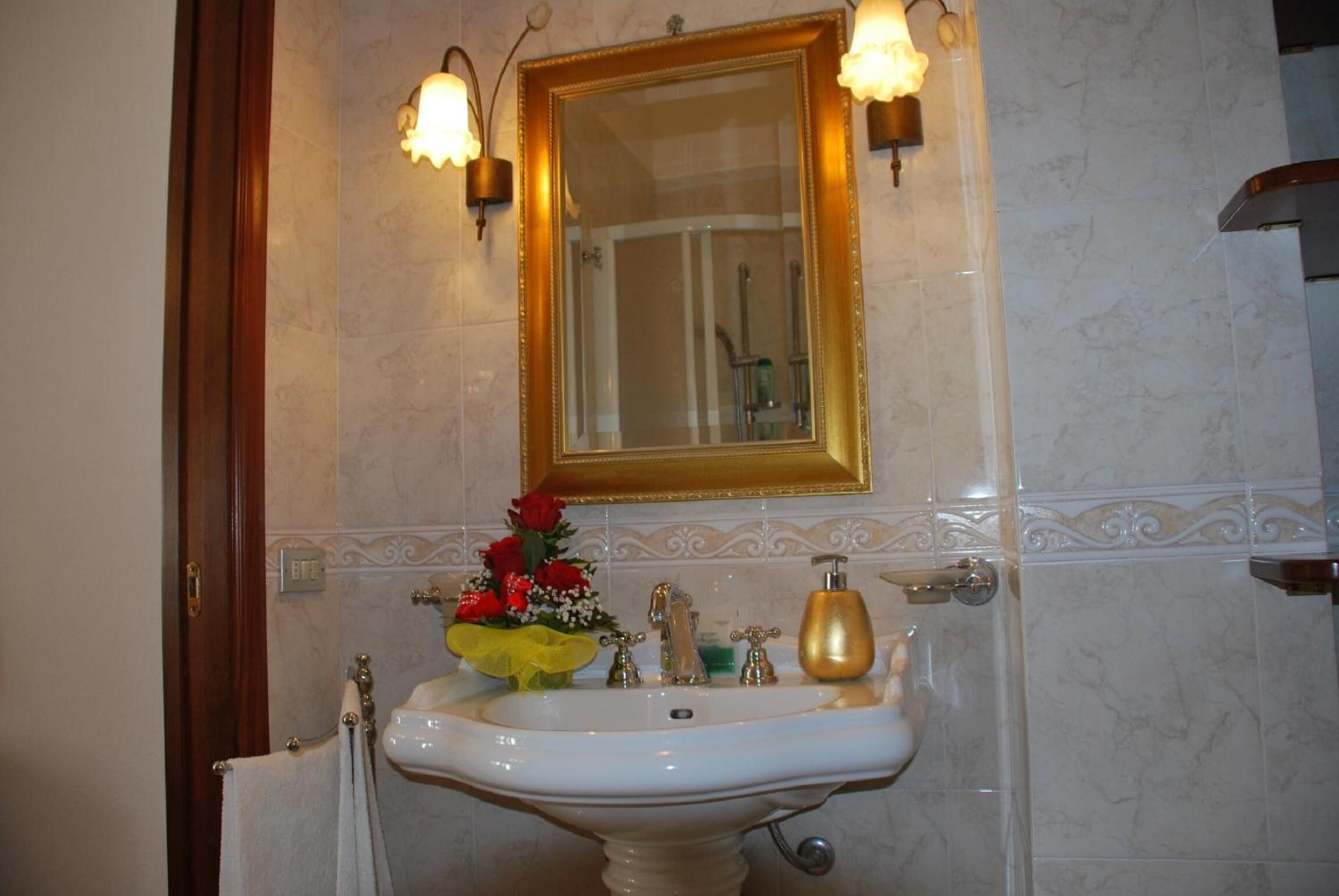 Un baño con lavamanos blanco, grifos dorados, dispensador de jabón dorado y un espejo con apliques de pared.