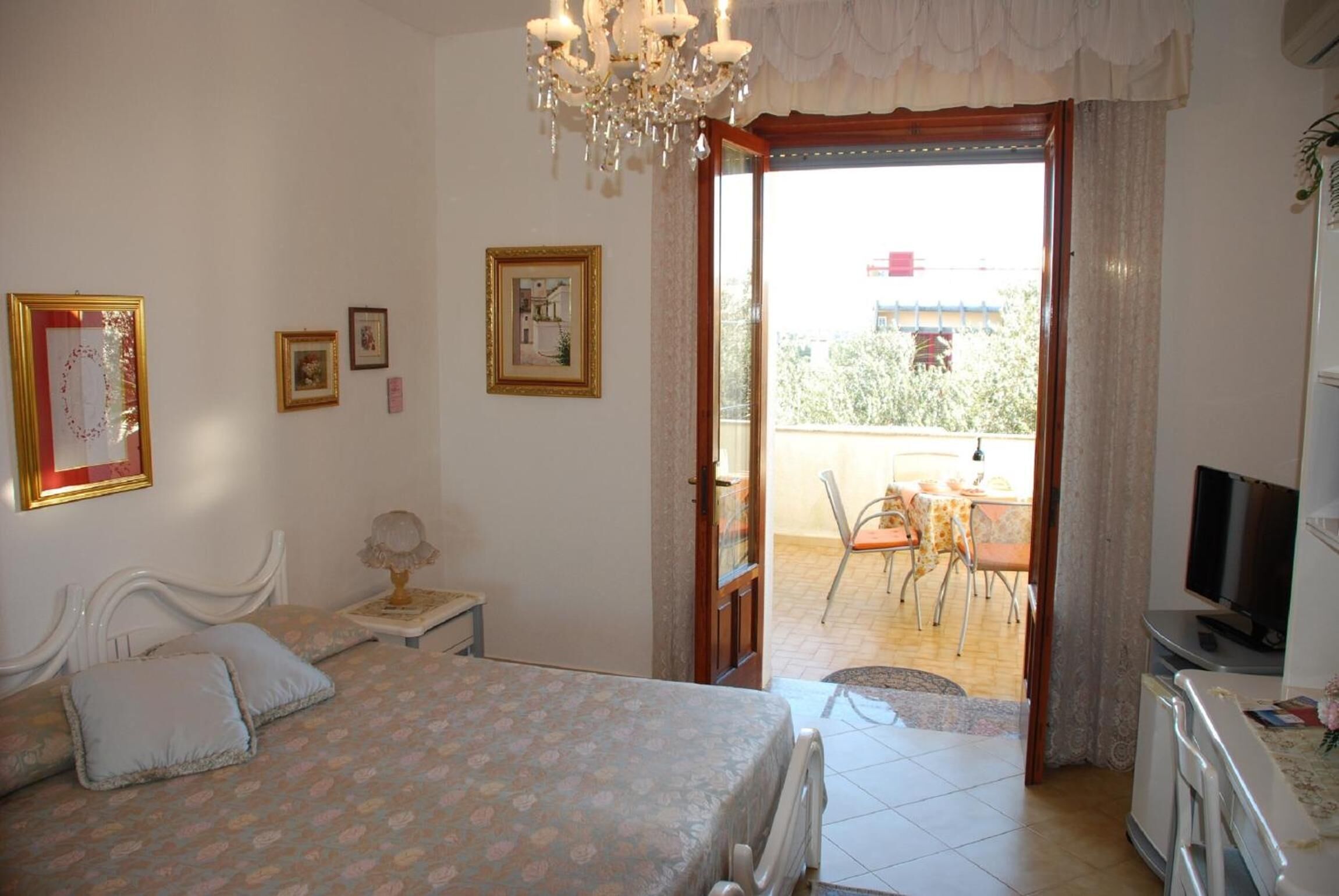 A bedroom with a bed, bedside table, lamp, and a TV. There is a balcony with a table and chairs.