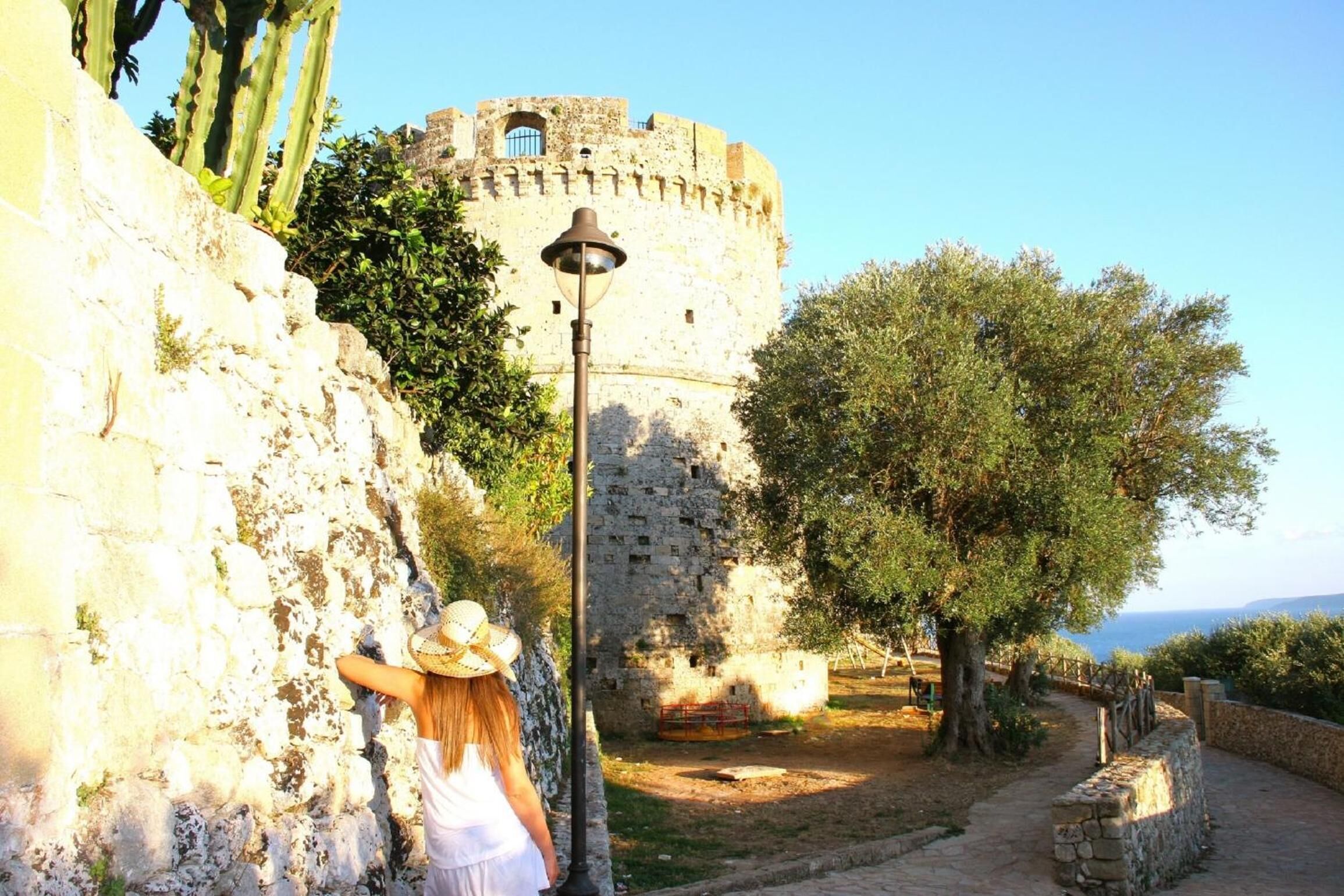 A historic stone wall with a person leaning against it, a lamp post, and a view of a castle-like structure in the background.