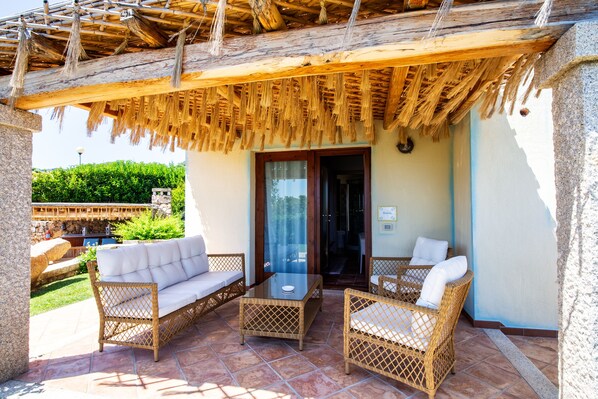 An outdoor patio with wicker furniture, a glass table, and a thatched roof. - Hotel Costa Caddu (San Teodoro)
