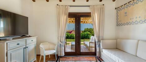 A room with a wooden ceiling, a TV on a cabinet, a sofa, a chair, a patterned rug, and a view through a glass door to an outdoor area with a table and chairs.