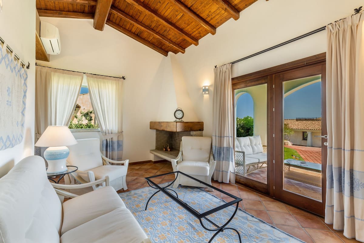 A spacious living area with a wooden ceiling, a fireplace, white furniture, and a view through a glass door to an outdoor patio.