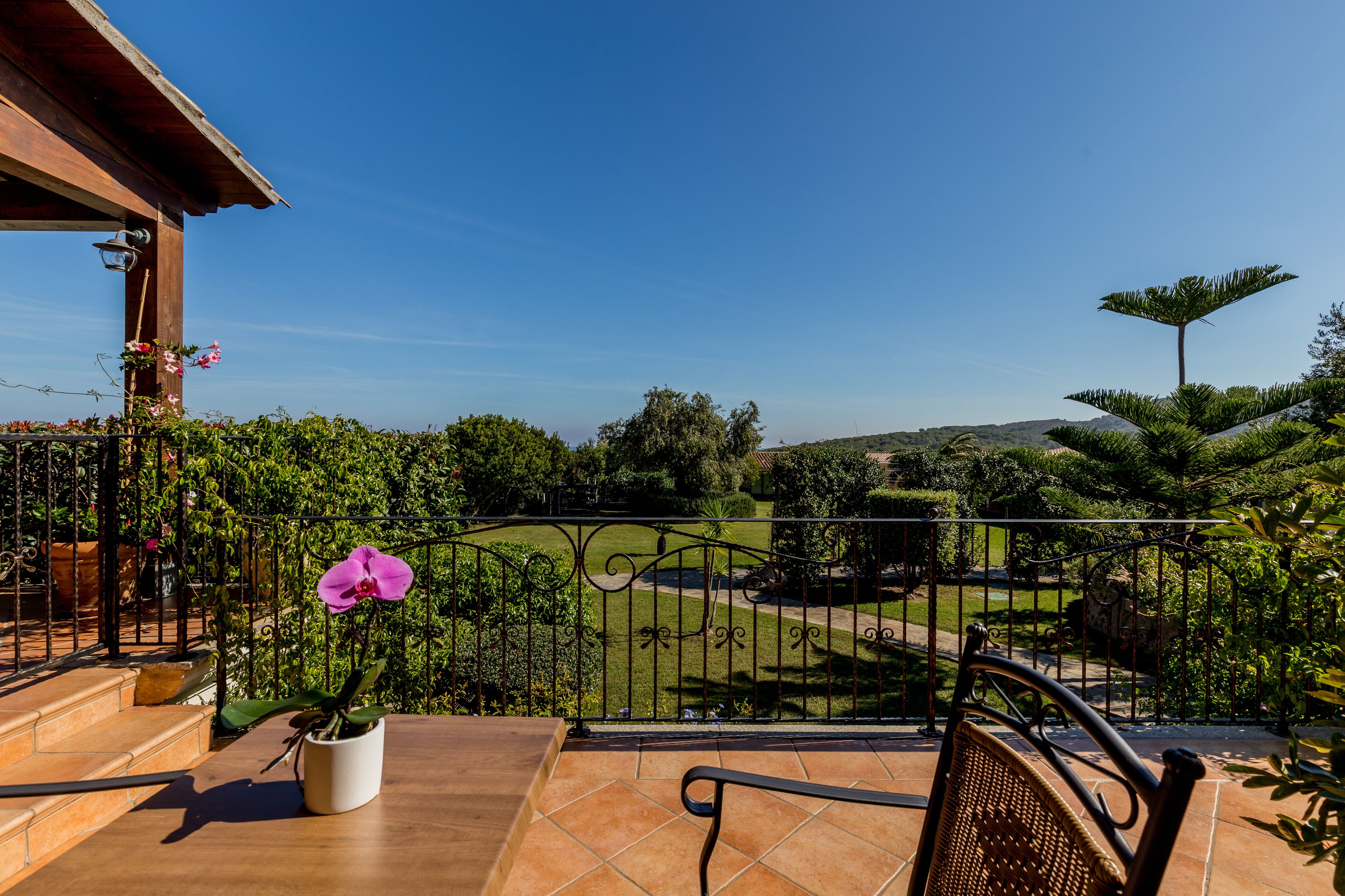 A balcony with a potted orchid, a wicker chair, and a view of a landscaped garden and distant hills.