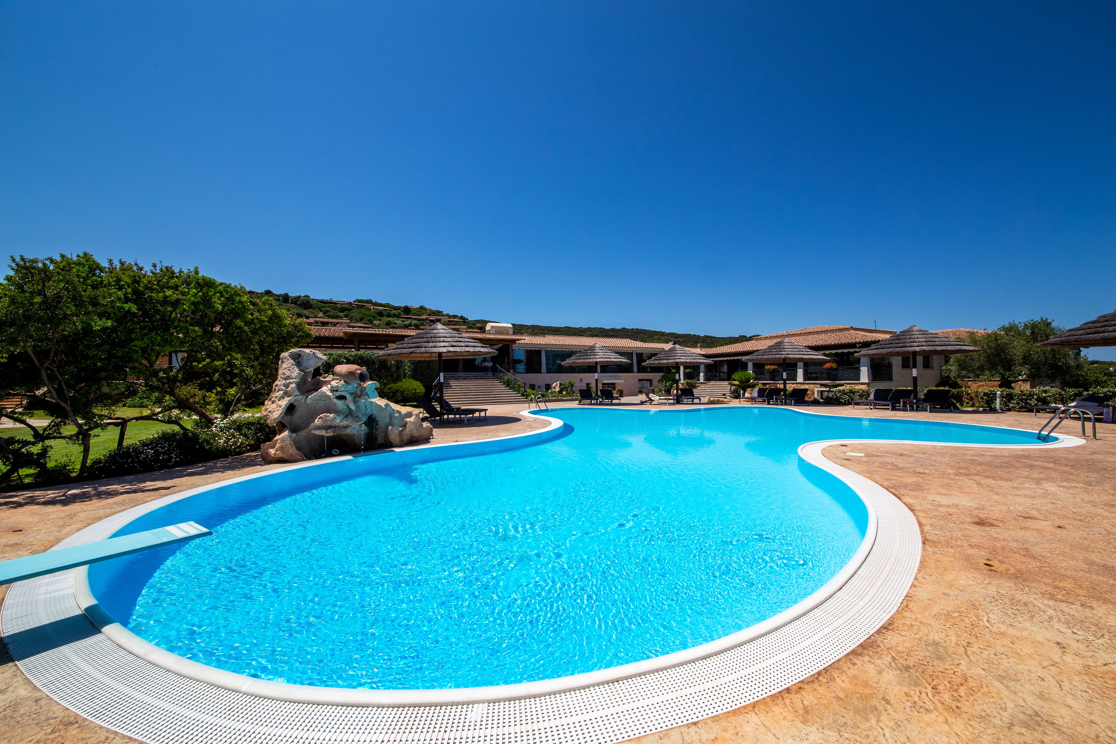 A large, curved swimming pool with a clear blue center, surrounded by a tiled deck and lounge chairs.