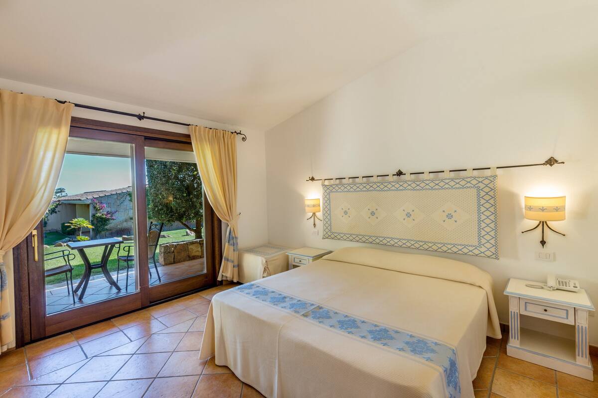 A hotel room with a bed, a small bedside table, a nightstand, a window with curtains, a view of the outside, and a wall-mounted lamp.
