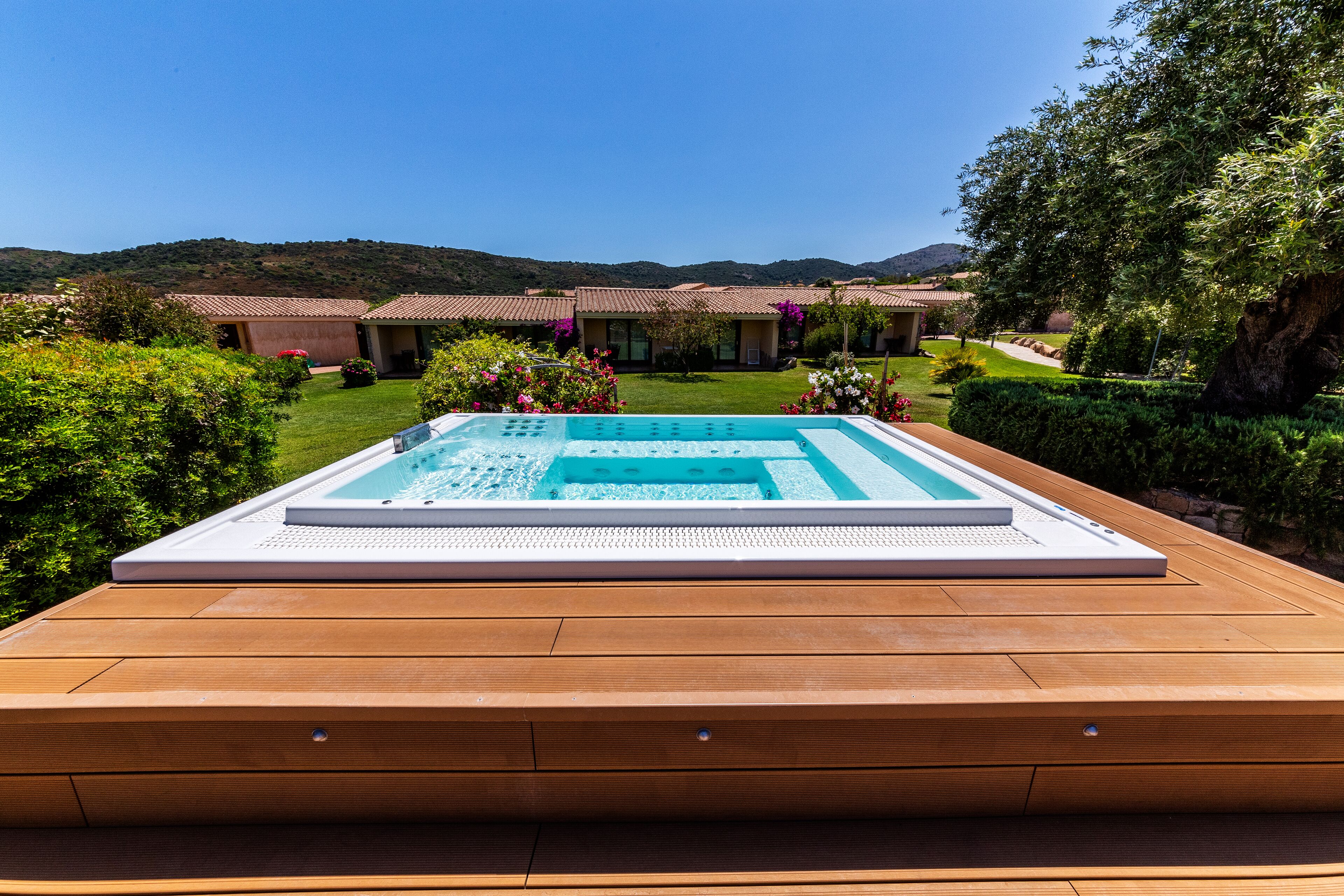 A wooden deck with a rectangular hot tub and a pool, surrounded by lush greenery and a view of a hillside.