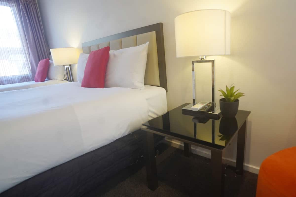 A hotel room with a bed, bedside lamps, a nightstand with a phone and a plant, and an orange chair.