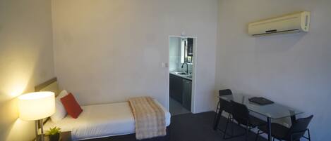 Habitación de hotel compacta con una cama individual, mesita de noche con lámpara, una pequeña planta en maceta, escritorio con silla y un equipo de aire acondicionado montado en la pared.