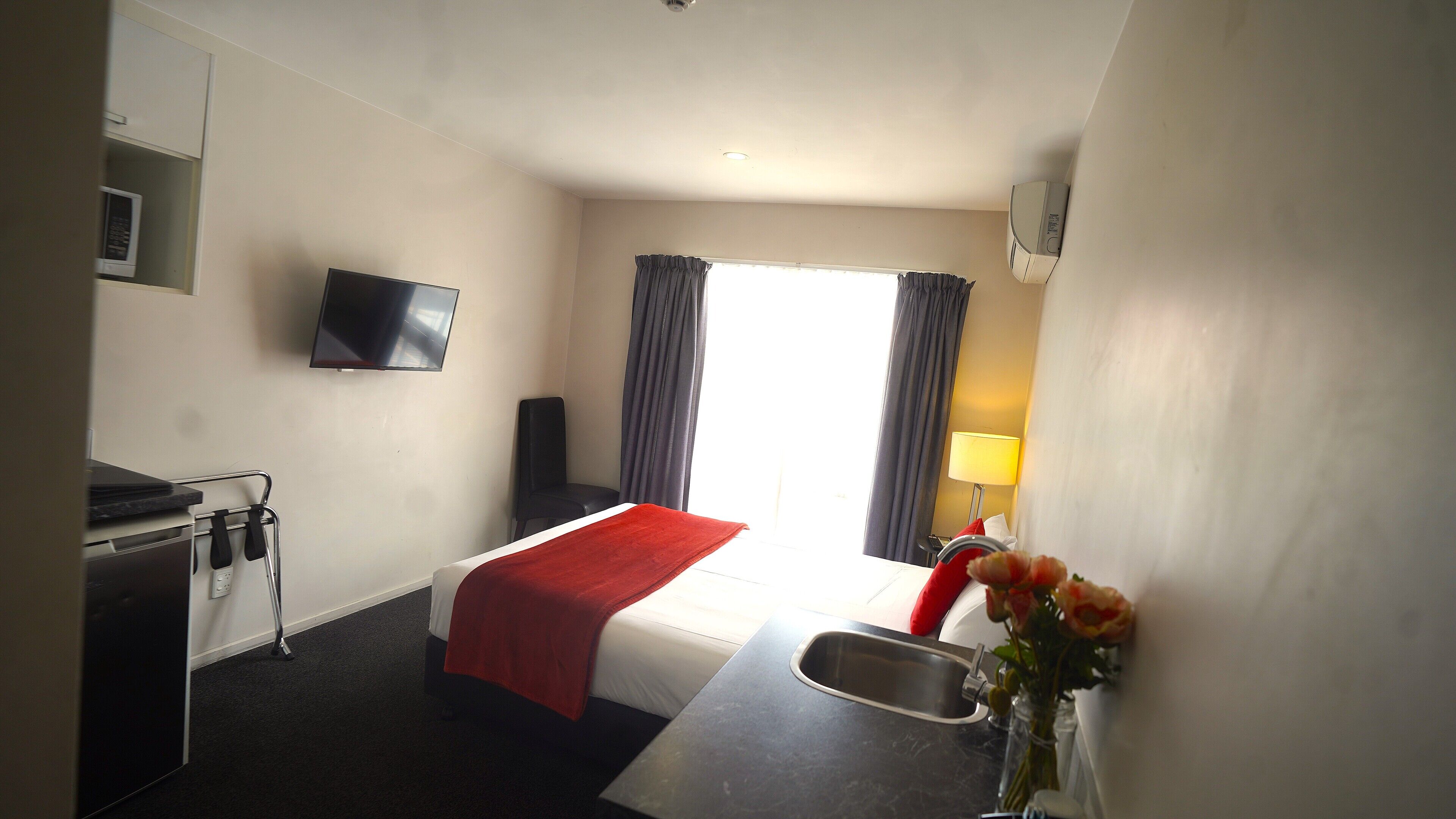 A hotel room with a bed, a TV mounted on the wall, a desk with a sink, and a window with curtains.