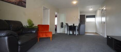 A living room with a black leather couch, an orange chair, and a dining area with a table and chairs. There is a kitchen area with a refrigerator and a window.