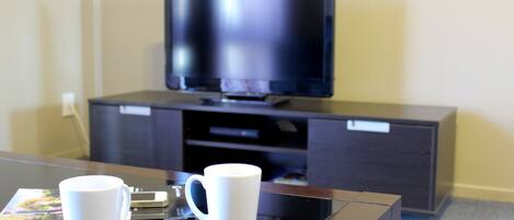 Habitación de hotel con una televisión de pantalla plana sobre un mueble de madera oscura, dos tazas blancas sobre una mesa de vidrio y un libro.
