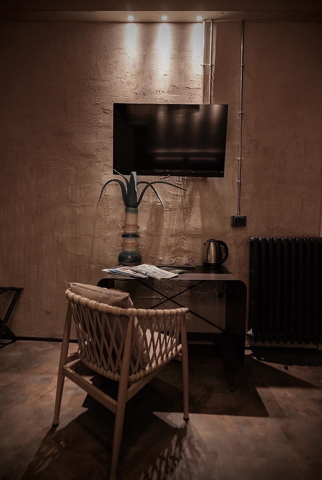Habitación con televisión de pantalla plana montada en la pared, una silla de mimbre, una mesita con hervidor y un radiador.