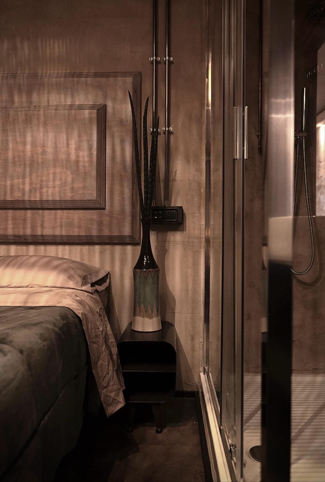 A bedroom with a bed, a nightstand, a vase, and a shower area.