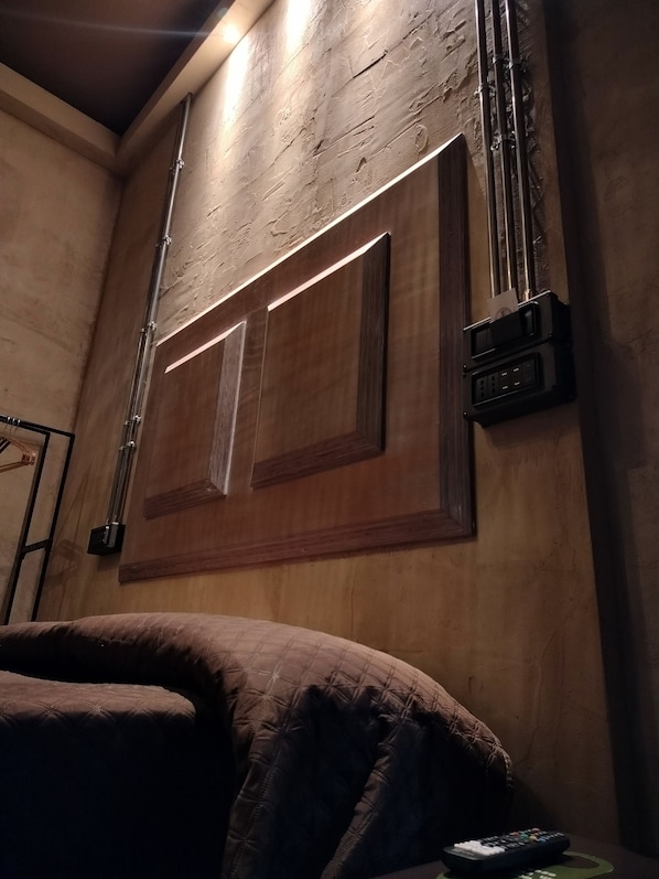 Una habitación con cabecera de madera, una cama, una lámpara fijada a la pared y un enchufe.