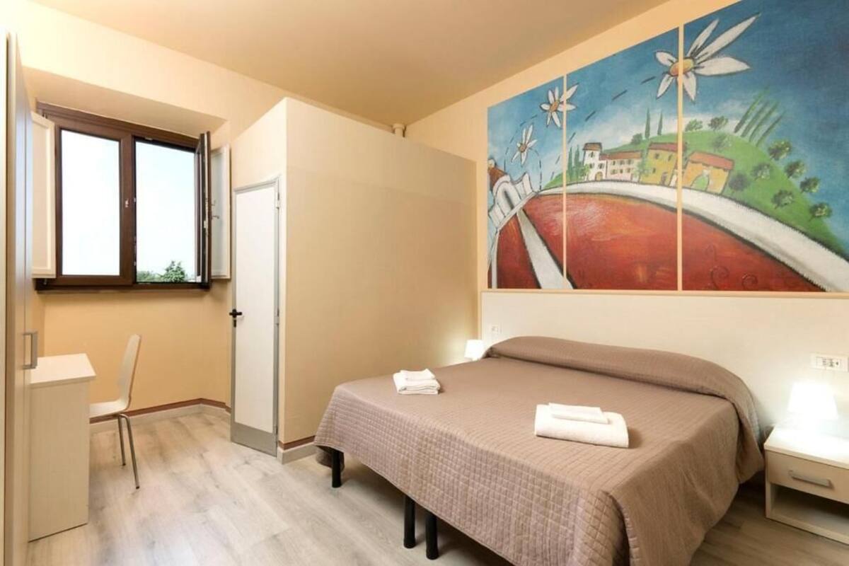 A bedroom with a bed, a desk, and a chair. There is a window and a painting on the wall.