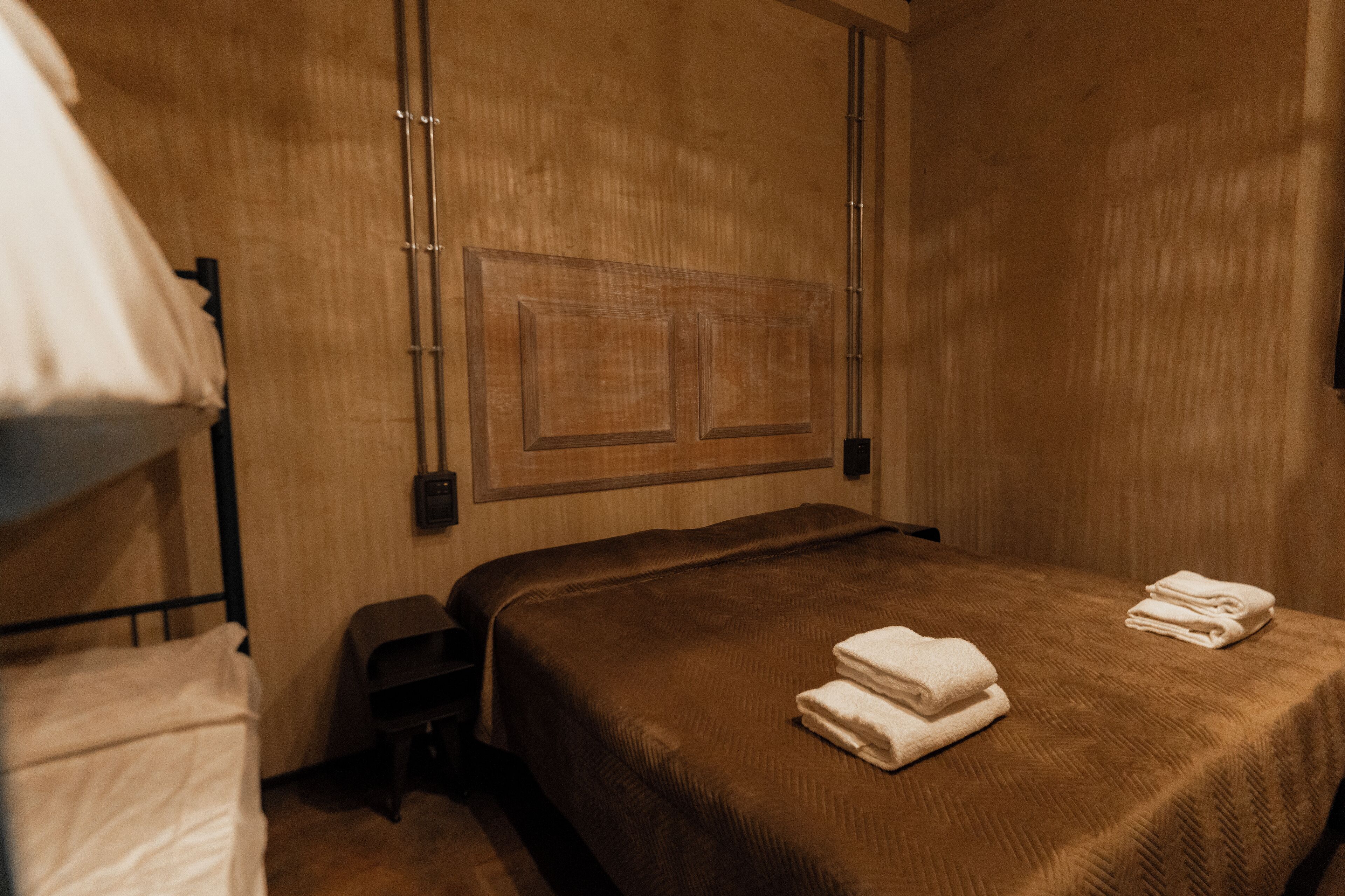 Camera d'albergo con una testiera in legno, un letto singolo con copriletto marrone e due asciugamani bianchi piegati con cura sul letto.