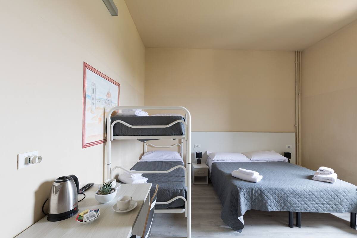 A hotel room with bunk beds, a desk with a kettle, a small plant, and a framed picture on the wall.