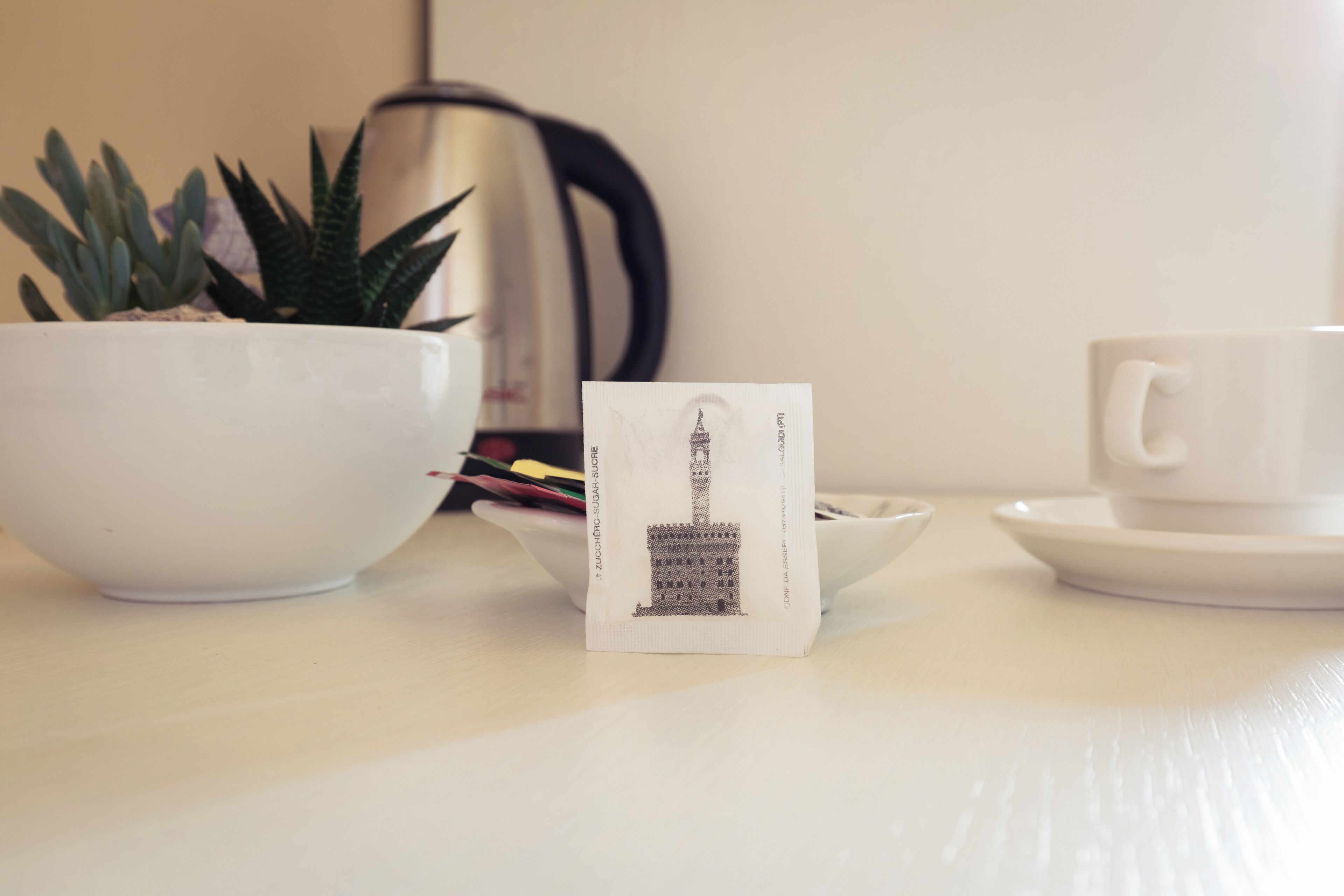 Een witte tafel met een witte kop, schotel en een kleine witte kom. Er staat een potplant en een theezakje op de tafel.