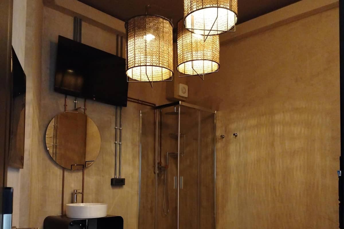 A hotel room with a bed, a round mirror, a sink, a shower, and two hanging lamps.