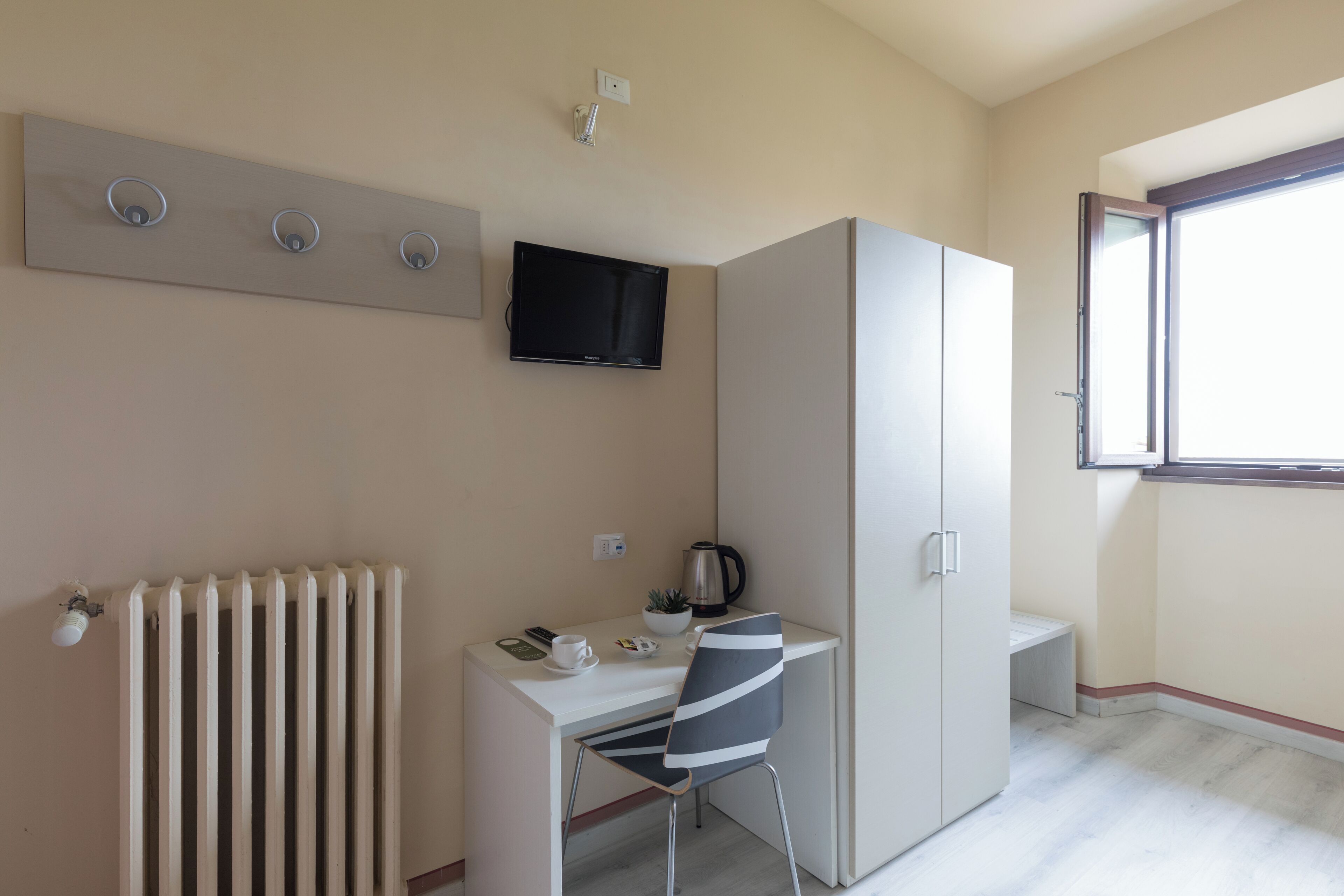 Um quarto pequeno e bem iluminado com uma escrivaninha branca, uma cadeira, um radiador, uma televisão fixada na parede e uma janela com vista para o exterior.