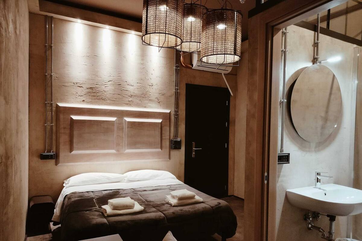A modern bedroom with a large bed, a bedside table, a lamp, and a bathroom with a sink and mirror.