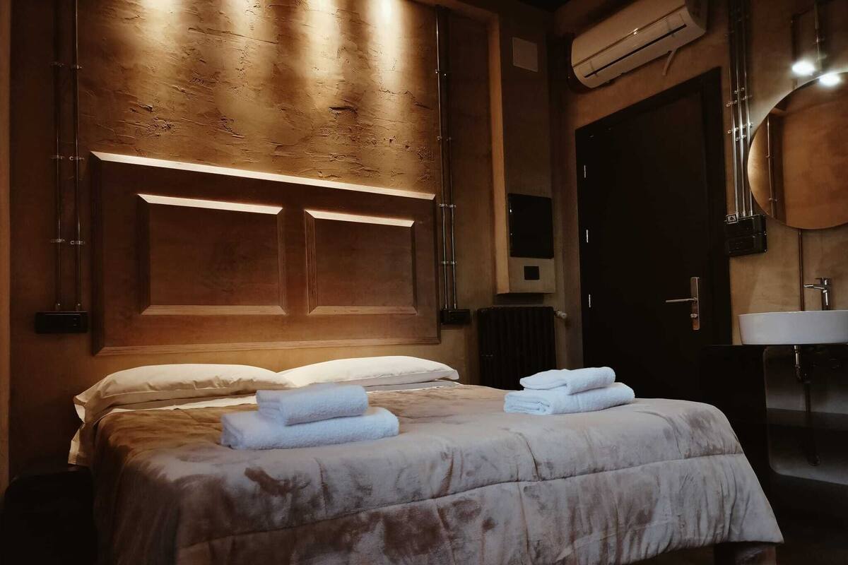 A hotel room with a large bed, a wooden headboard, a bedside table, and a bathroom with a sink and mirror.