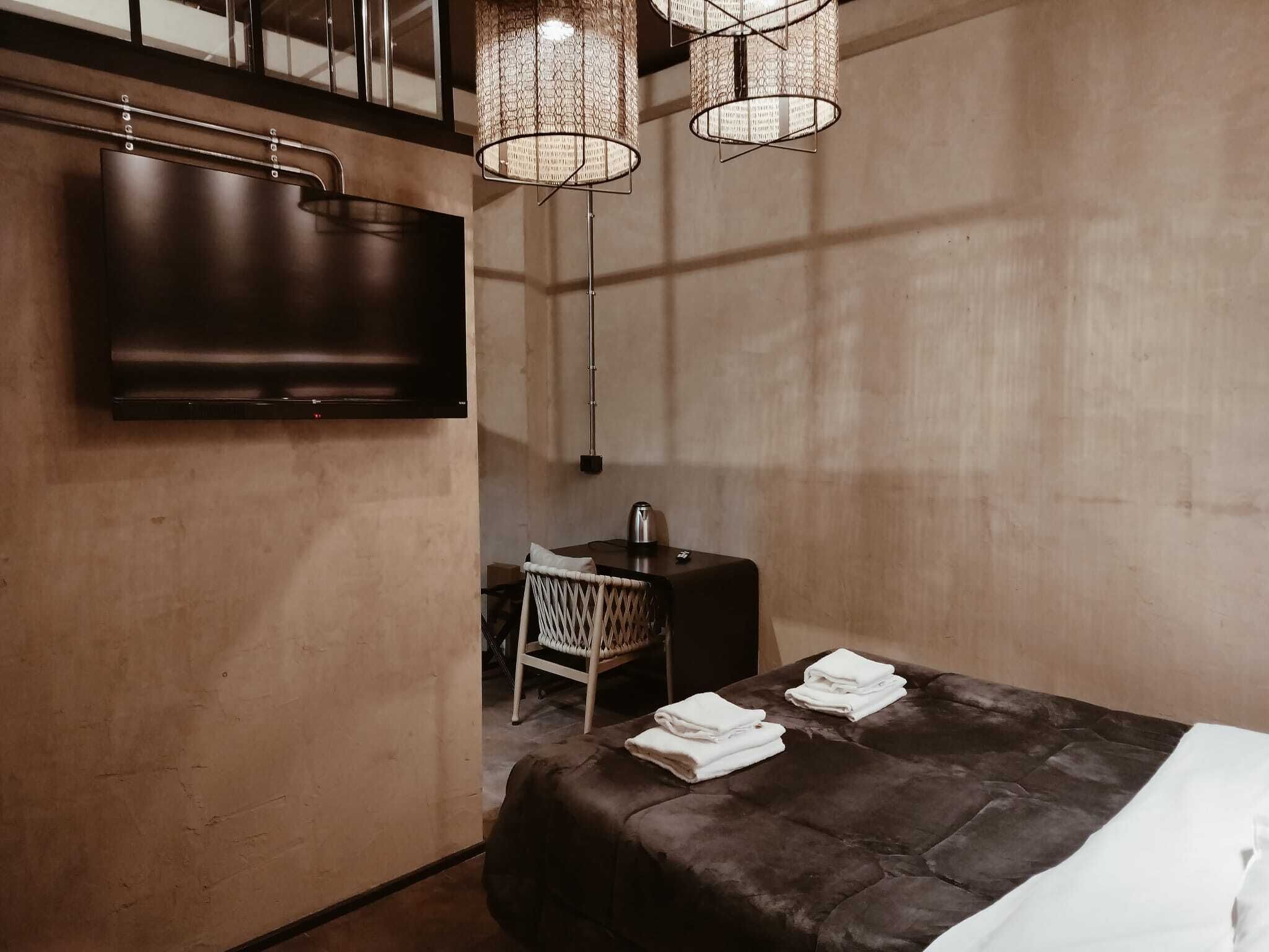 A modern hotel room with a large bed, a flat-screen TV, a small table with a chair, and a hanging light fixture.