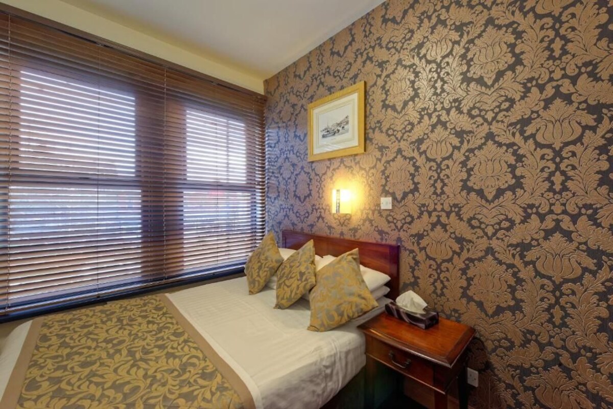 A hotel room with a bed, a nightstand, a framed picture on the wall, and a window with blinds.