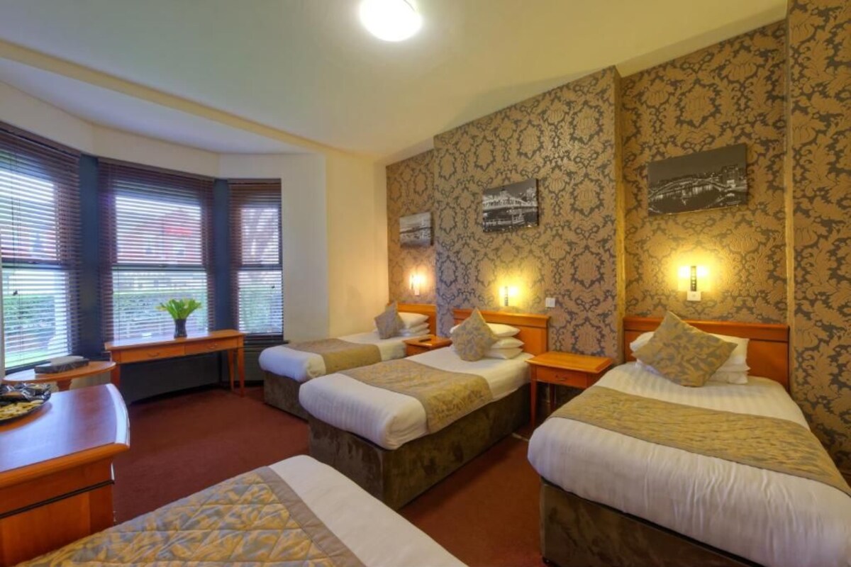 A hotel room with three single beds, a desk, and a chair. There are two windows with blinds, a small table with a plant, and wall-mounted lamps.