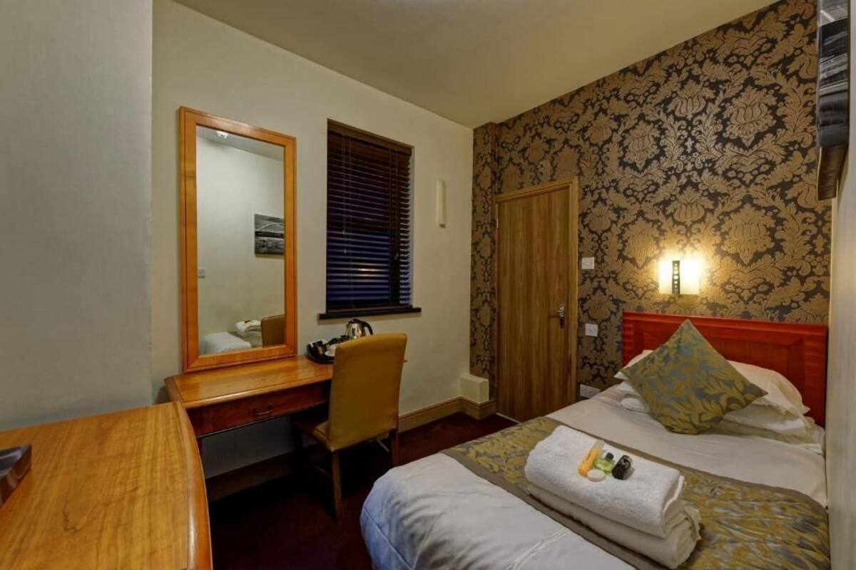 Une chambre d’hôtel avec un bureau en bois, une chaise, un lit avec une tête de lit rouge et un papier peint à motifs.