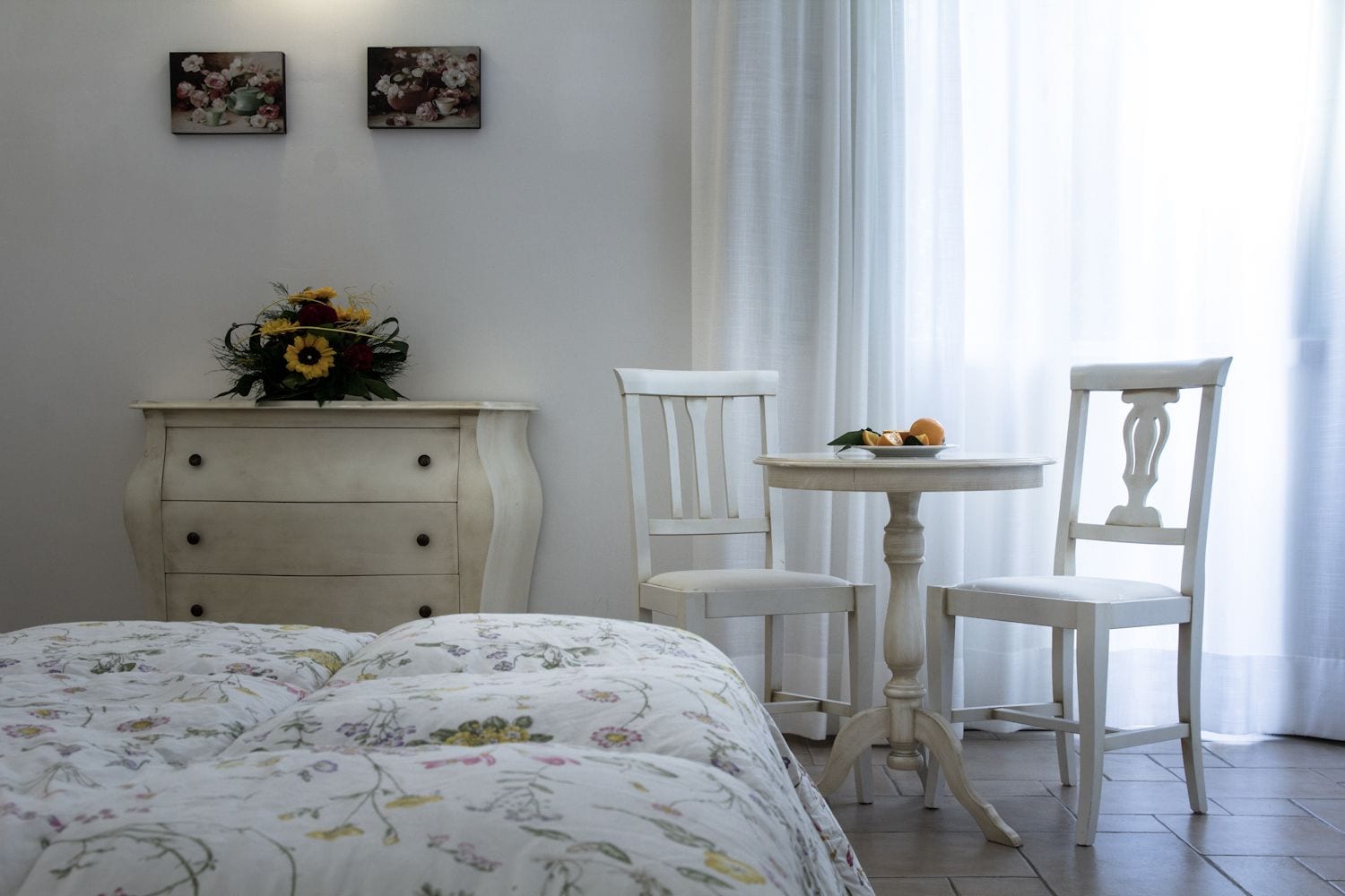 A bedroom with a bed, a dresser, a chair, a table, and two wall-mounted paintings.